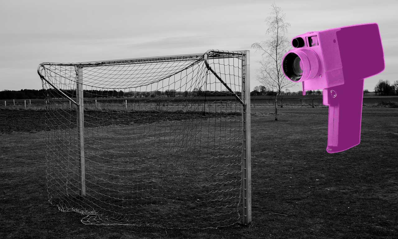 Schwarz-weiß Bild eines Fuballtors auf einer wiese. Bildrechts eine alte Handkamera, die auf das Tor gerichtet ist.